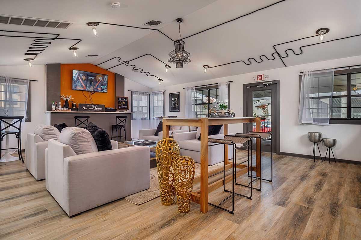 A modern living space featuring a cozy seating area with white sofas, a wooden dining table, and decorative gold sculptures. Unique black wiring decorates the ceiling, and large windows provide natural light. A bar area is visible in the background, adding to the inviting atmosphere.