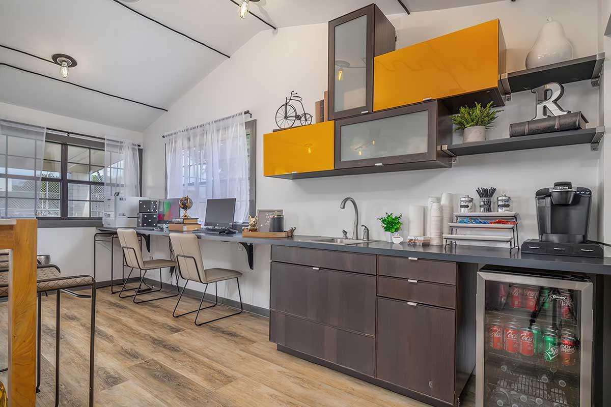 Modern kitchen with a sleek dark cabinet featuring bright orange accents. Includes a sink, coffee maker, and shelves with decorative items. Two workstations with computers are visible in the background. The space is well-lit with large windows, showcasing wood flooring and a cozy seating area.