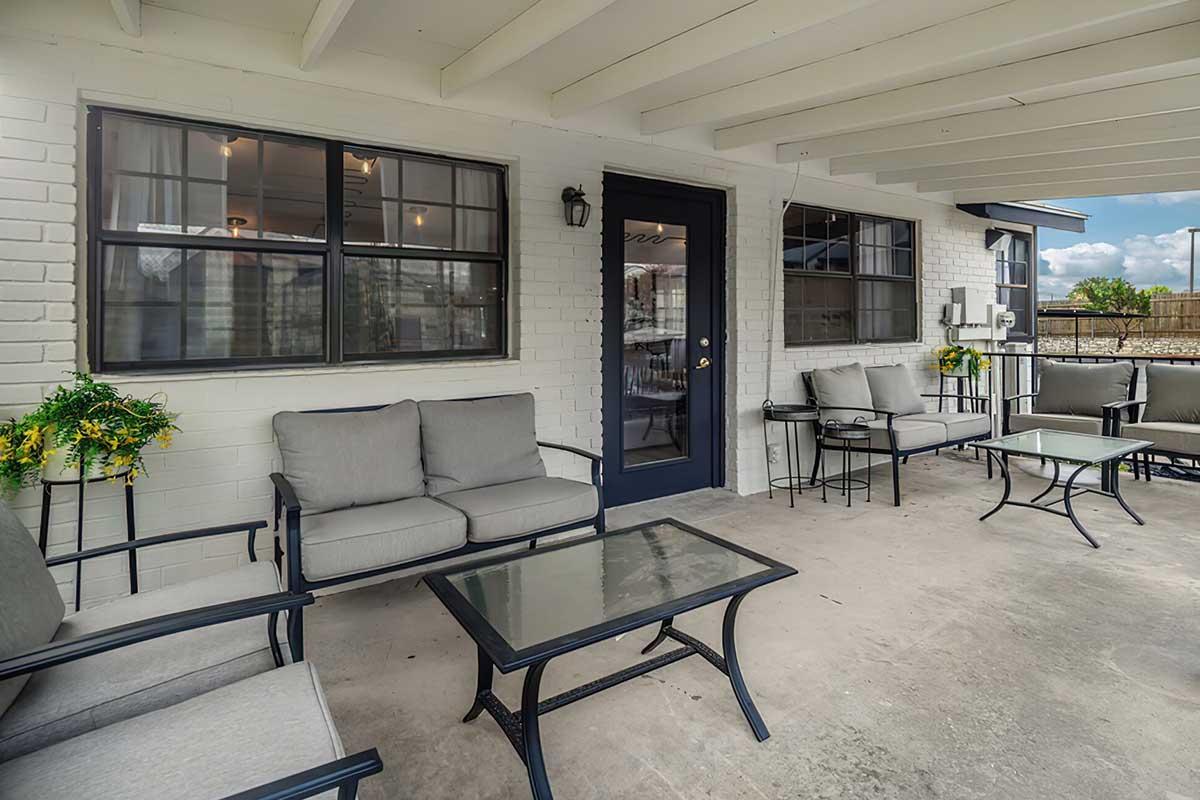 A cozy patio area featuring a seating arrangement with gray cushions, a glass-top table, and decorative plants. The space is partially covered, with walls painted in white. Large windows provide natural light, and a door leads to another part of the building. The atmosphere is inviting and relaxing.