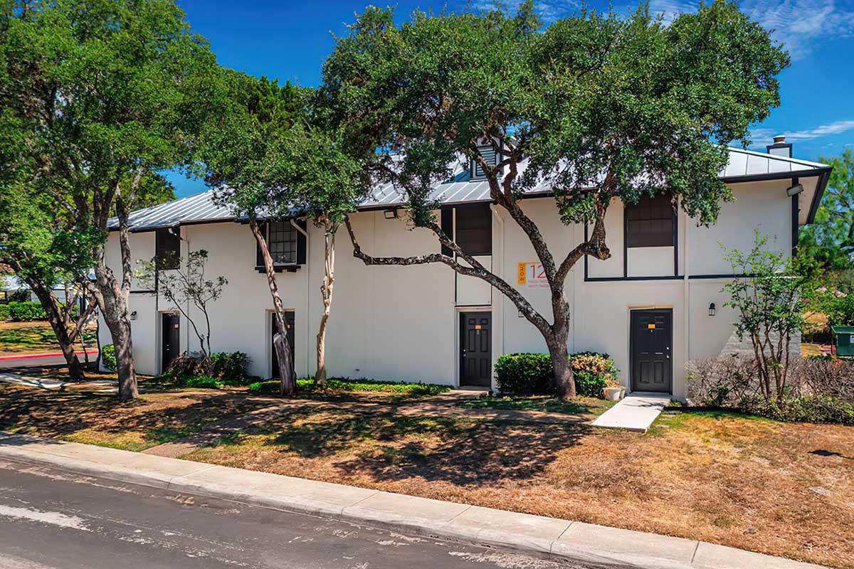 A two-story building with a light-colored façade and a metal roof, surrounded by trees and shrubs. The front features four doors, two on each level, and a walkway leading to the entrance. The area is well-maintained with a grassy patch and a paved road in front. Bright blue sky enhances the overall appearance.