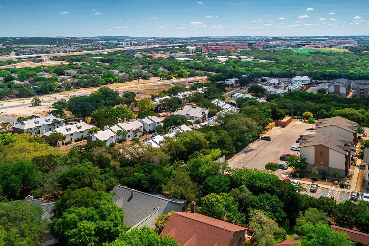 Aerial view of a suburban landscape showcasing a mix of residential buildings and greenery. The scene features a variety of homes surrounded by trees, with some open land and distant urban development visible in the background under a clear blue sky.
