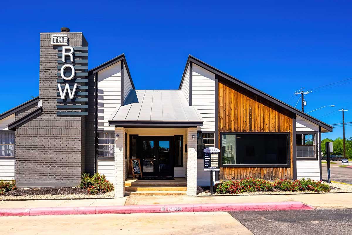 Exterior view of a modern building featuring a combination of gray and wooden siding. The entrance has large windows and a welcoming door under a peaked roof. A sign on the left side reads "THE ROW." The surrounding area is landscaped with small plants and has a clear blue sky above.