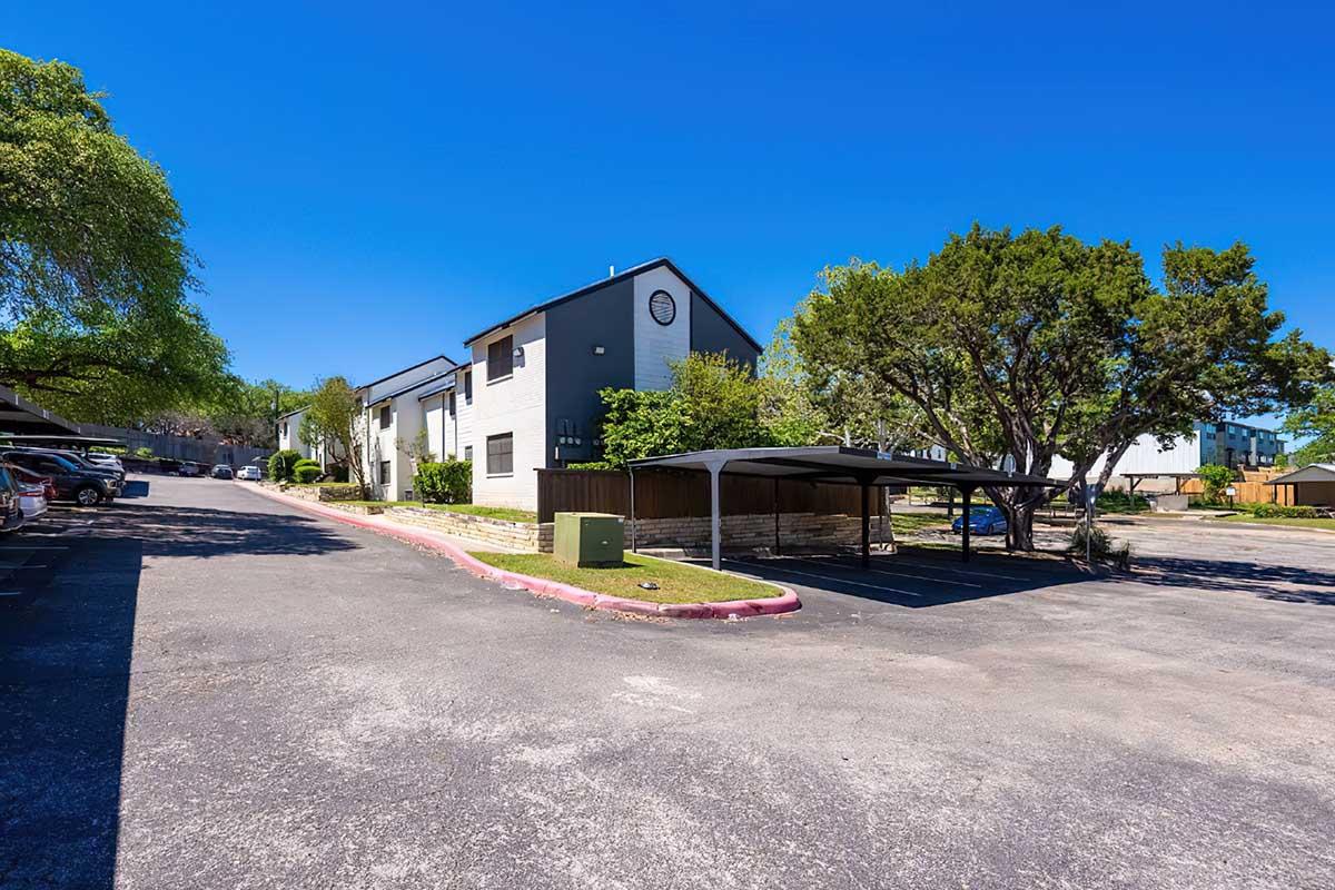 A sunny day view of a residential area with two-story apartment buildings on the left, surrounded by greenery. A partially shaded carport is visible in the foreground, along with several parked cars along the driveway. Blue sky and sparse clouds complete the scene.