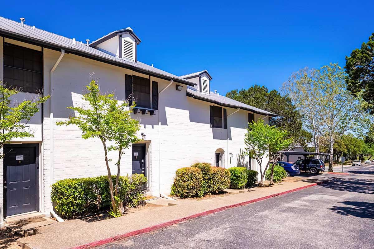 A modern two-story apartment building with a light-colored exterior and gray doors. The walkway is lined with small shrubs and trees, and there are parked cars nearby. The sky is clear and blue, indicating a bright day.