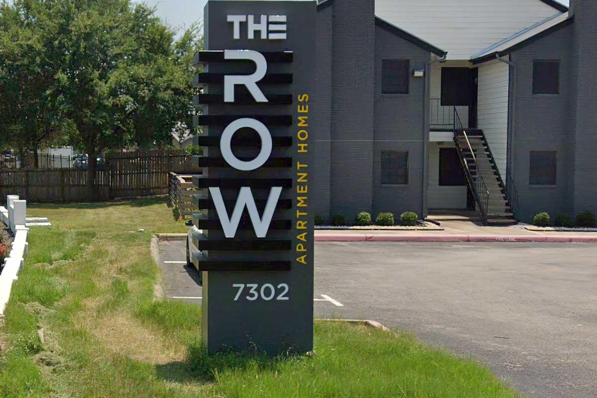 Sign for The Row Apartment Homes, featuring the name "THE ROW" in large letters, with "APARTMENT HOMES" in smaller text below. The sign is located at 7302, set against a backdrop of a well-maintained apartment building and landscaped area.