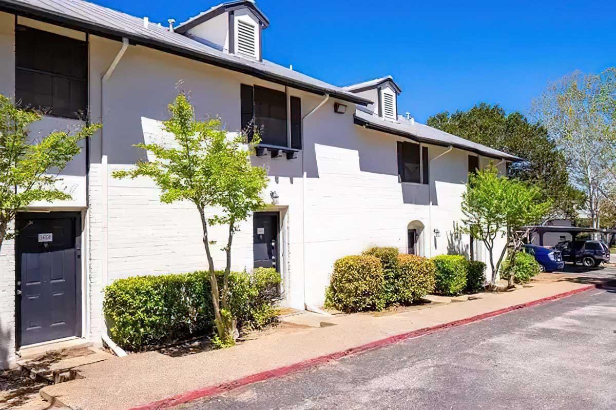 A two-story apartment building with a white exterior, featuring multiple entrances and small trees along the walkway. The building is set against a clear blue sky and includes shrubs near the entrance. A parking area is visible in the background.