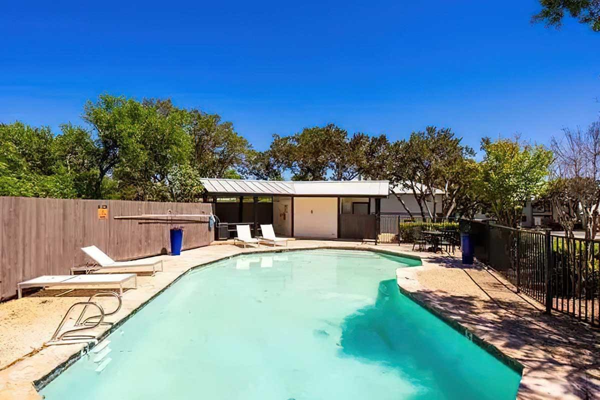 A clear swimming pool surrounded by lounge chairs, with a wooden fence and trees in the background. The sky is bright blue, and there is a modern building partially visible behind the pool area. The setting is serene and inviting, ideal for relaxation.
