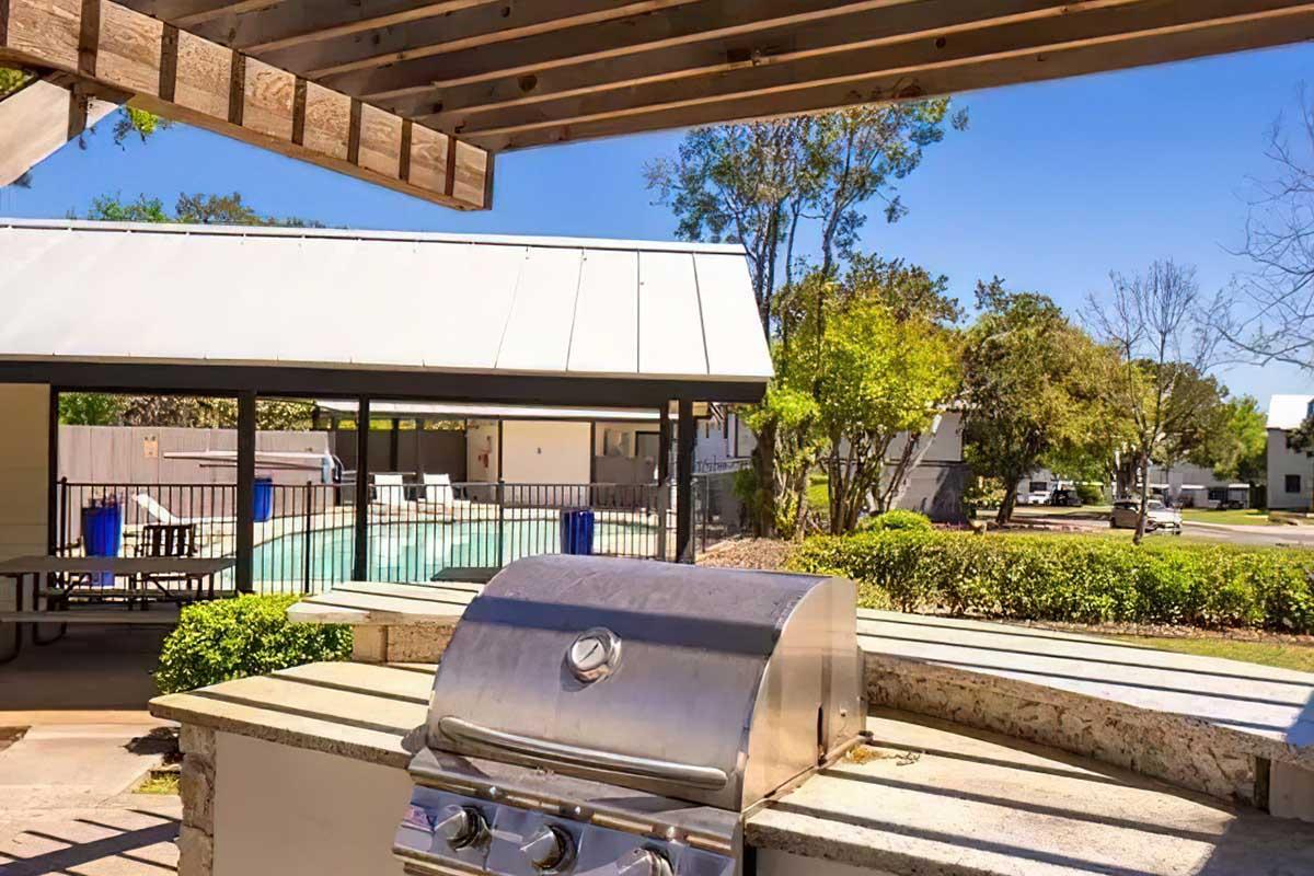 Outdoor grilling area with a stainless steel barbecue grill, surrounded by greenery. In the background, a swimming pool is visible, enclosed by a fence, and there are picnic tables under a shaded structure. Bright blue sky and trees provide a pleasant atmosphere.