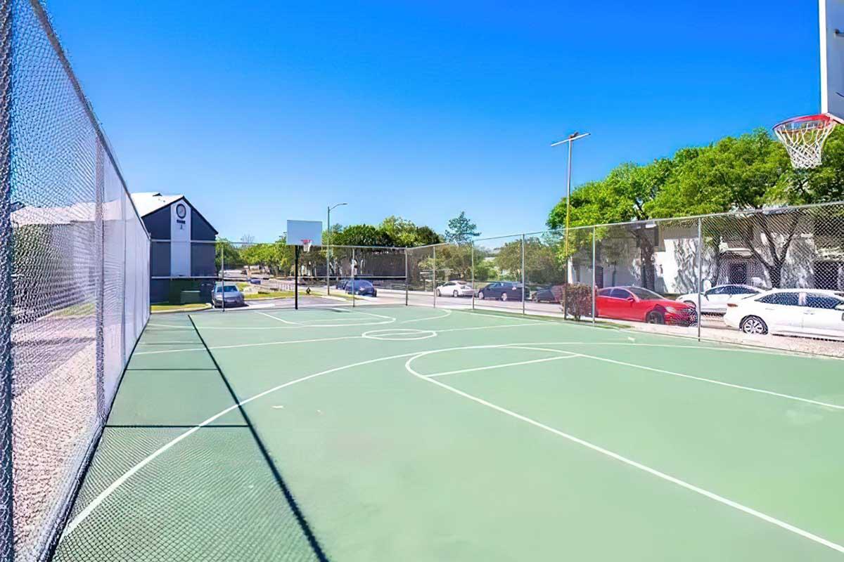 A view of a fenced outdoor basketball court featuring two hoops, with painted lines for play. In the background, there are parked cars and a building surrounded by trees under a clear blue sky. The court appears well-maintained and inviting for recreational use.