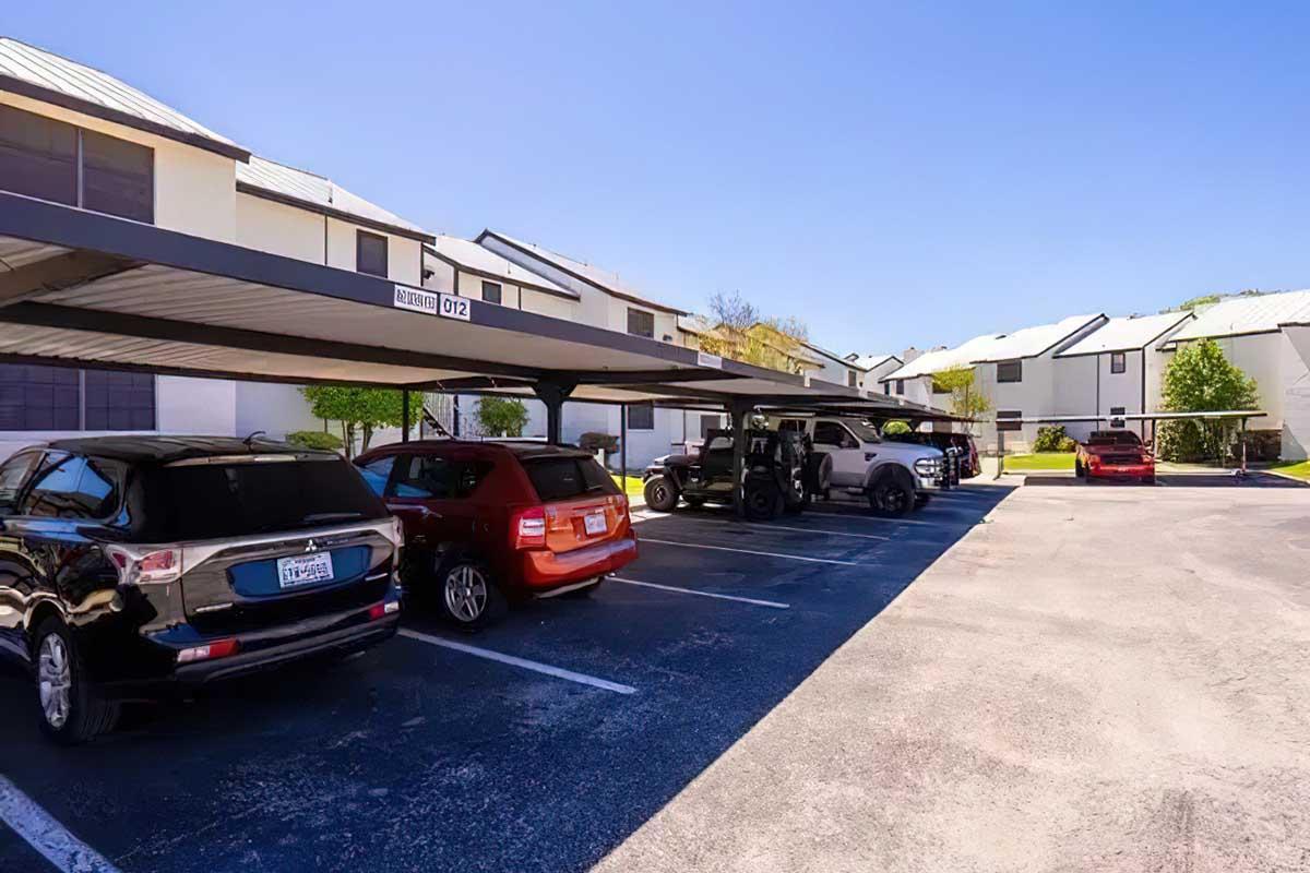 A sunny parking lot with covered spaces, featuring several parked vehicles including a black SUV, a red car, and a gray truck. In the background, there are residential buildings with white walls and metal roofs. The scene is clear and well-lit, with a blue sky overhead.