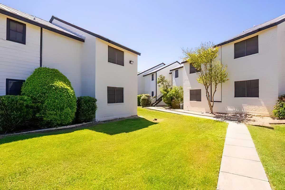 A well-maintained outdoor area featuring two white apartment buildings with black windows, surrounded by neatly trimmed greenery and a pathway. The scene is bright and sunny, highlighting the manicured lawn and landscaping between the buildings.