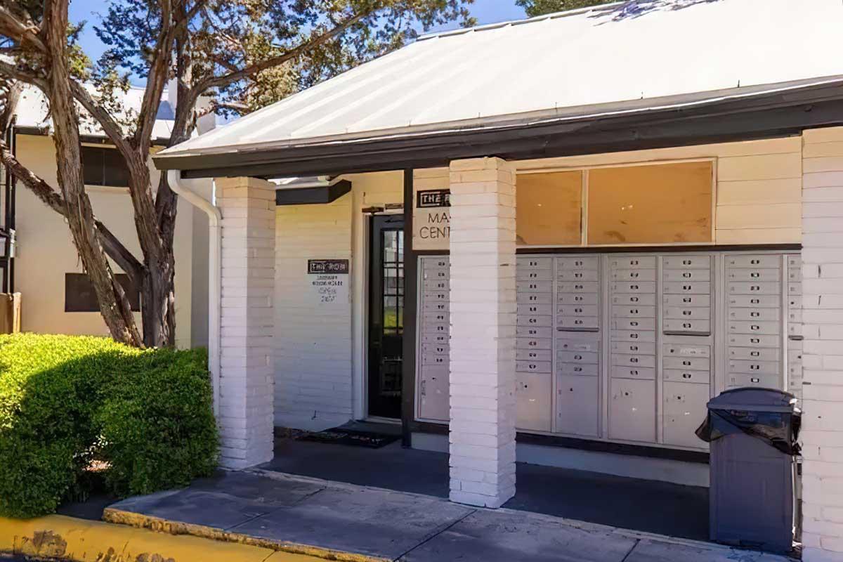 A small mail center building with white brick columns and a sloped roof. There are multiple mailboxes mounted on the wall, and a black trash can near the entrance. Green shrubs flank the sidewalk leading to the door, which has a sign indicating the center's name. The windows are covered, giving a quiet appearance.