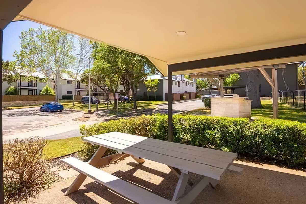 A shaded picnic table under a structure, surrounded by neatly trimmed bushes. In the background, a sunny street with a few parked cars and residential buildings is visible, along with trees lining the area, creating a peaceful outdoor setting.