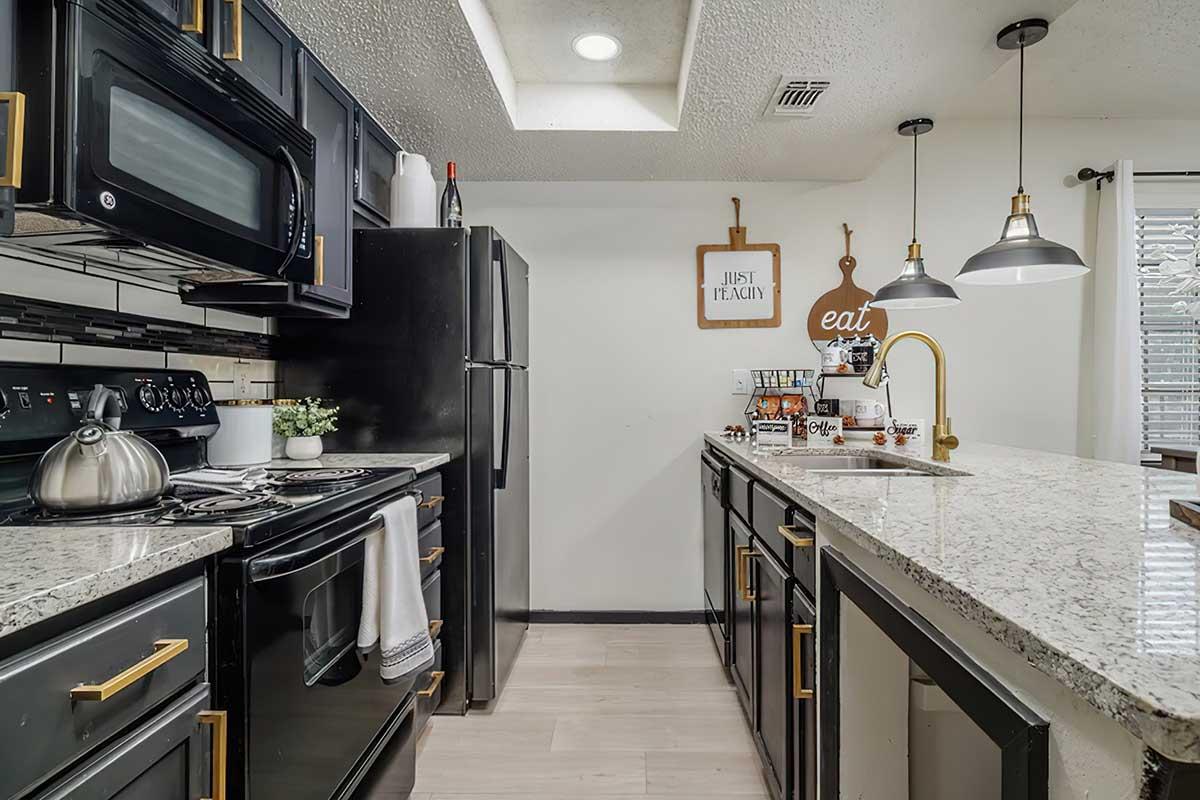 Modern kitchen featuring black appliances, a granite countertop, and sleek cabinetry. The space includes a gas stove, microwave, and an island with seating. Decorative items like a kettle and a plant add a cozy touch, while pendant lights illuminate the area under a skylight. Wall decor includes a wooden sign with "Just Be You" and "Eat."