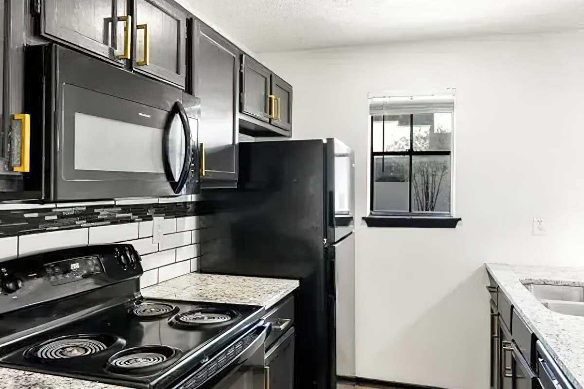 Modern kitchen featuring black cabinetry, a white and black tiled backsplash, granite countertops, and stainless steel appliances, including a microwave and refrigerator. The space is well-lit with a window allowing natural light, giving a bright and inviting atmosphere.