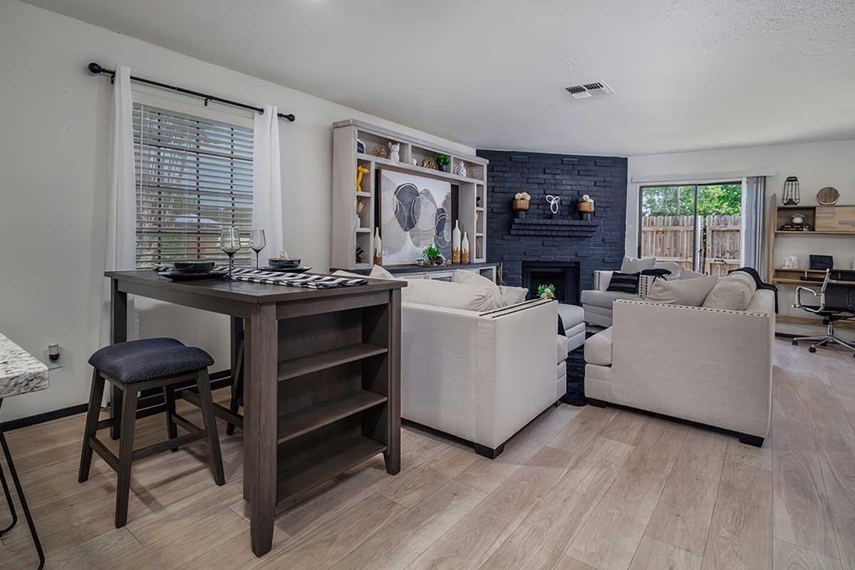 A cozy living room featuring a modern design with a black stone fireplace, beige sofas, and a small dining table. Large windows allow natural light in, and decorative shelving adds character. The room has light hardwood flooring and a mix of functional and decorative elements, creating an inviting atmosphere.