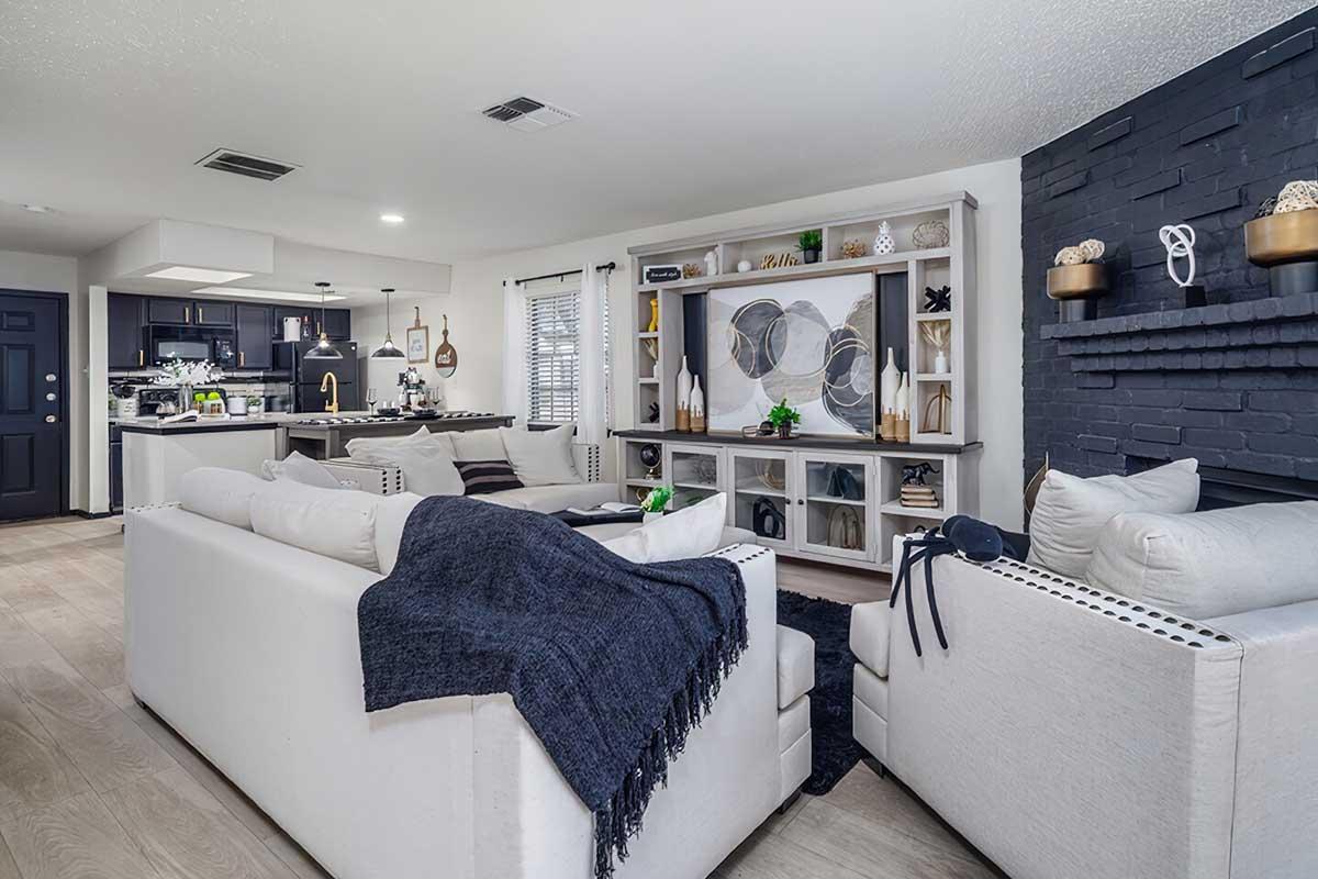 A modern living room featuring a white sectional sofa with a dark throw blanket, a stylish coffee table, and a large entertainment unit with decor items. The walls are painted light colors, and there's a dark accent wall with decorative elements. The background shows a glimpse of a kitchen area.