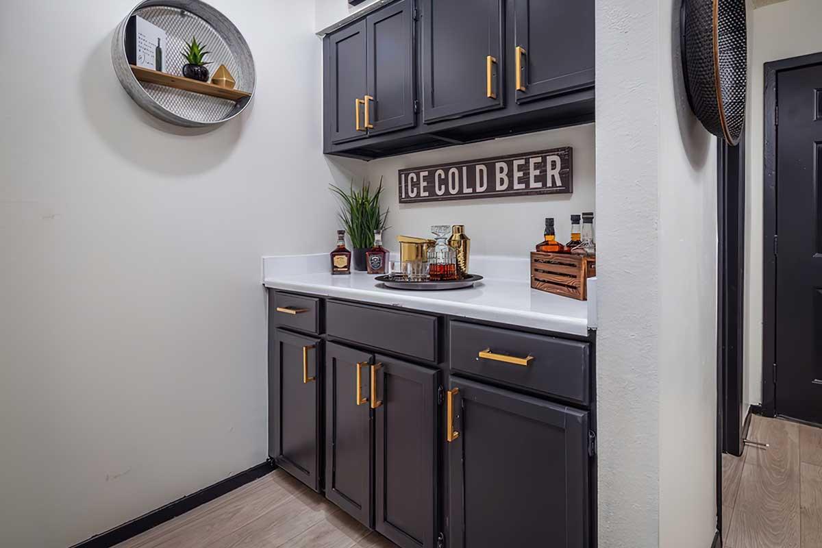 A stylish corner bar with dark cabinetry, a white countertop, and decorative items. It features a sign reading "ICE COLD BEER," a tray with beverage accessories, a wooden crate holding liquor bottles, and a circular shelf with small plants, creating a cozy and inviting atmosphere.