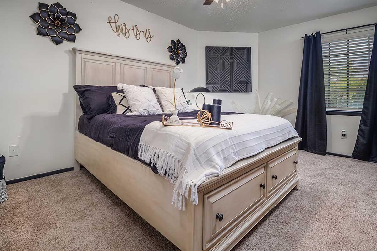 Cozy bedroom featuring a light wood bed frame with a white and navy bedding set. Decorative pillows are arranged on the bed, accompanied by a tray with decor items. The room has a neutral color palette with wall art, a window with blinds, and a plush carpeted floor, creating a warm and inviting atmosphere.