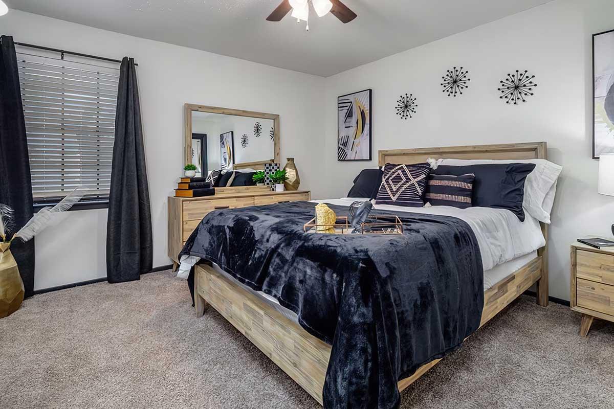 A cozy bedroom featuring a wooden bed with dark bedding, a decorative throw, and stylish pillows. A brown dresser with decorative items stands nearby, along with a mirror. The room includes a window with blinds, dark curtains, and modern wall art, creating a warm and inviting atmosphere.