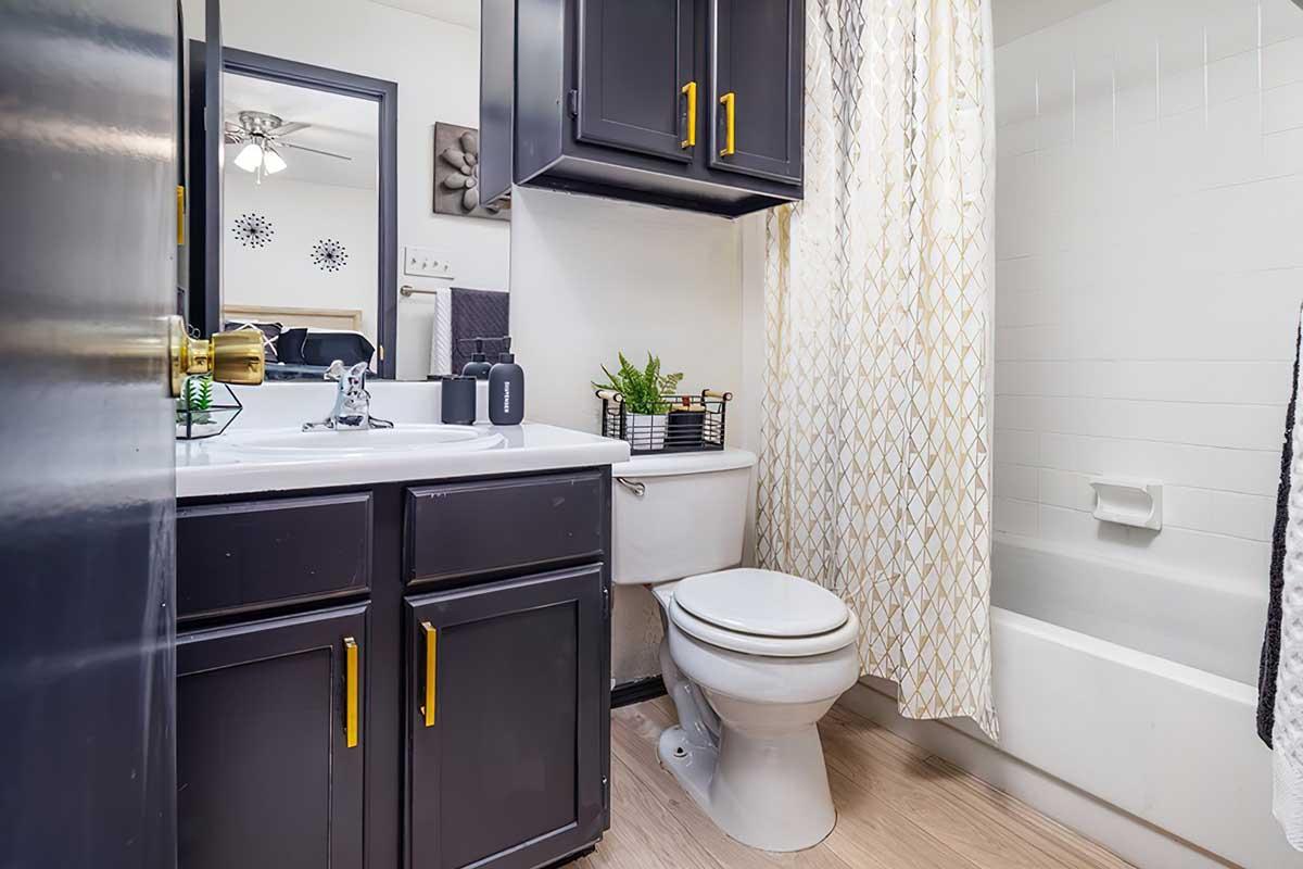 A small bathroom featuring dark cabinetry, a white sink, and a toilet. The bathtub has a white curtain with a geometric pattern. A small potted plant decorates the countertop. A ceiling fan is visible, and the overall decor has a modern, clean look with light wood flooring.