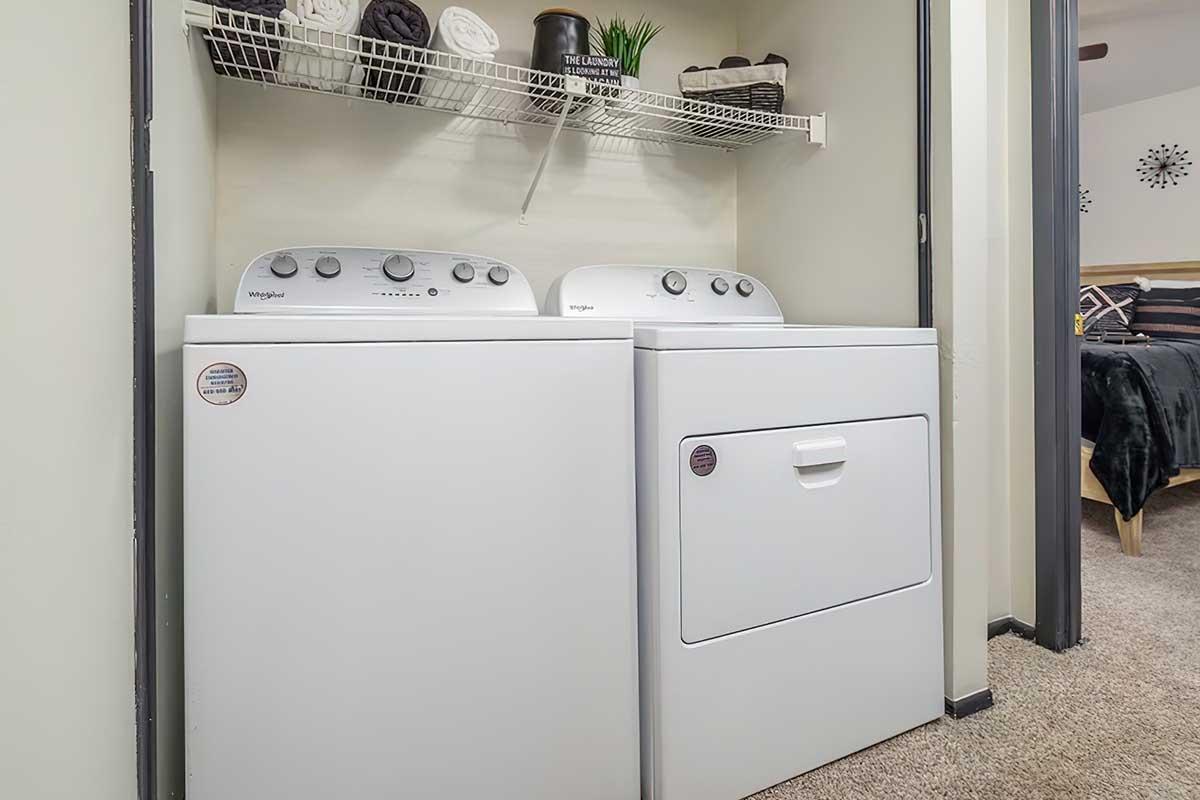 A washing machine and dryer stacked next to each other in a small laundry space. Above them, there is a shelf with neatly folded towels, a decorative plant, and a few storage baskets. The floor is carpeted, and a doorway leads to another room in the background.