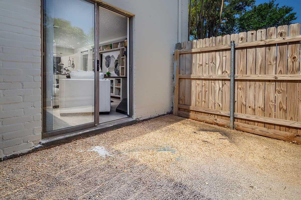 A small outdoor space featuring a gravel and dirt floor, with a wooden fence on one side. There is a glass sliding door leading to an interior room, partially visible through the opening. The area appears clean and well-kept, with greenery visible beyond the fence.