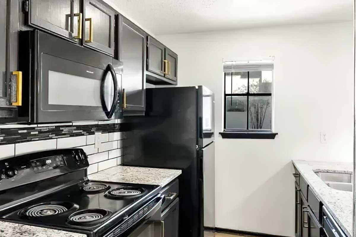A modern kitchen featuring black cabinets, a stainless steel microwave, and an electric stove. There's a black refrigerator and a window with a view outside. The countertops are made of granite, and the backsplash consists of black and white tiles, creating a sleek and stylish look.