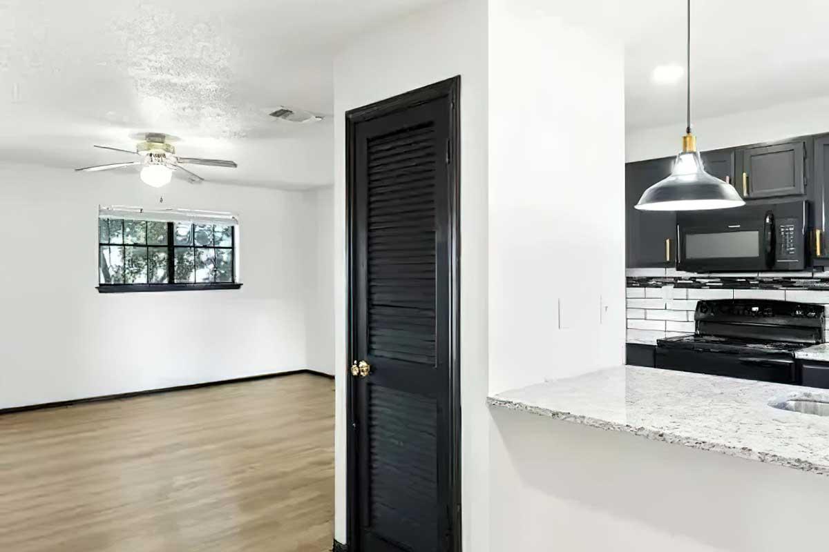 A spacious, well-lit interior with a view of a kitchen area featuring dark cabinetry, a black stove, and a granite countertop. To the left, there's a living area with wooden flooring and a ceiling fan, while a closet door is visible on the right, adding to the modern, open layout.