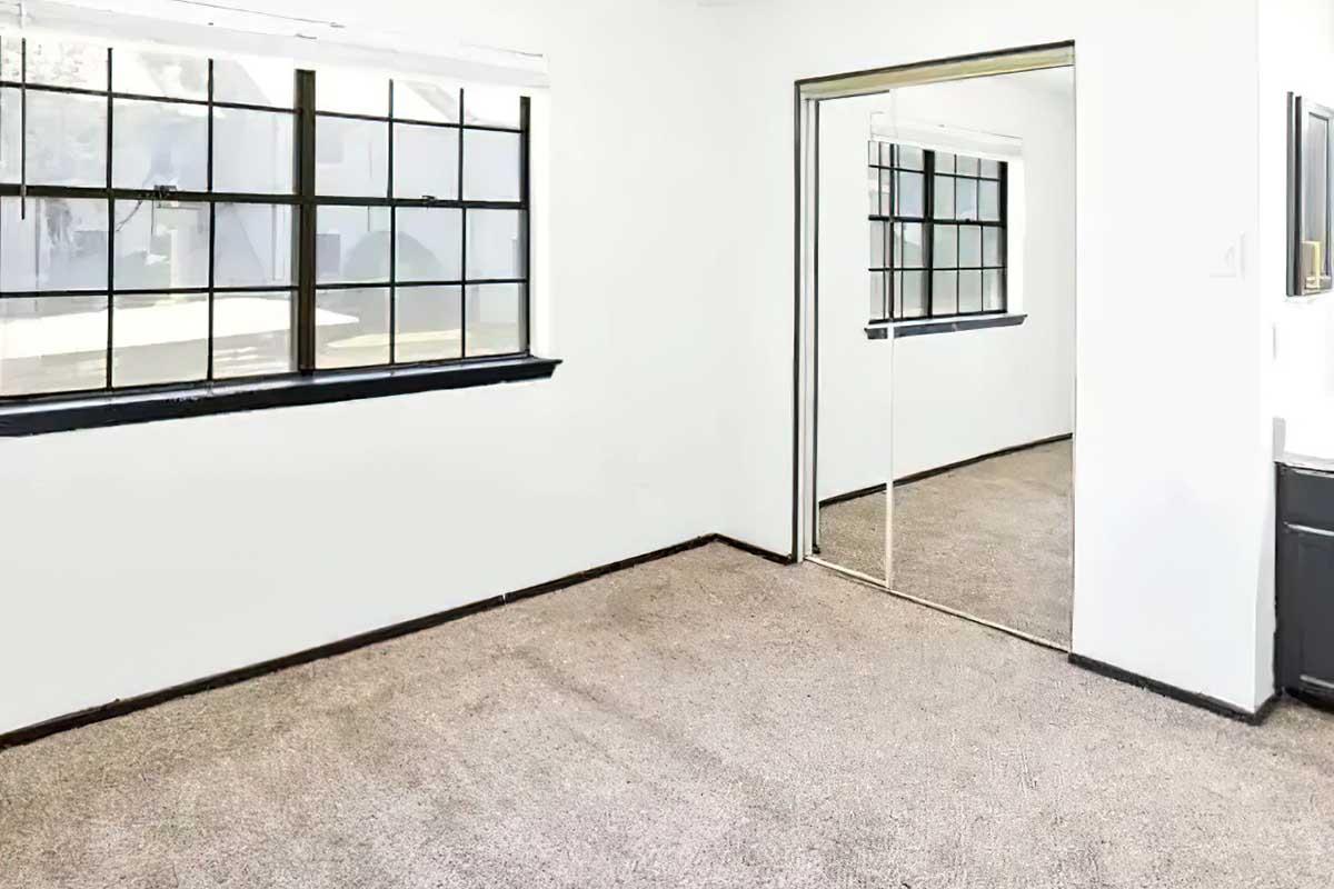 A vacant room with beige carpet, white walls, and large windows with black frames. One wall features a mirrored closet door reflecting the room. The space appears bright and uncluttered, suitable for various purposes.