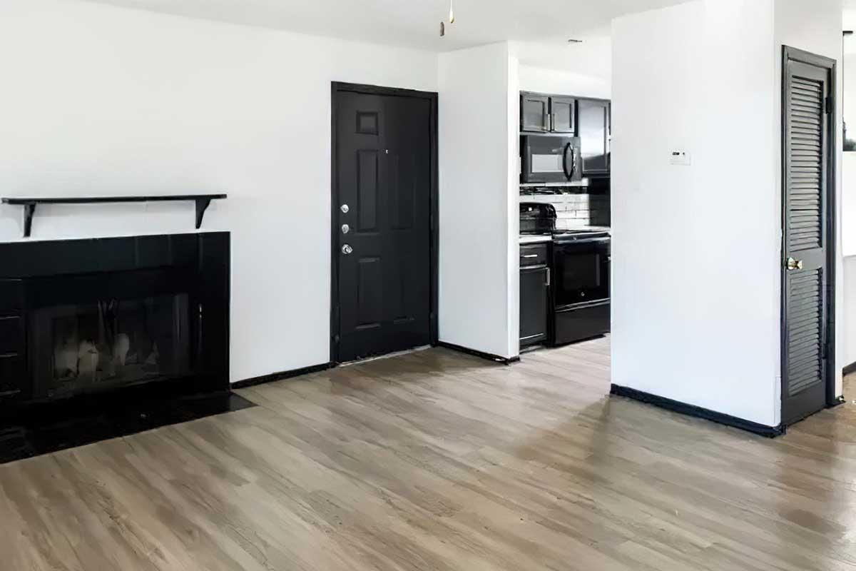 A spacious living room with light wood flooring, featuring a black fireplace on the left and a black front door. A doorway leads to a modern kitchen with black appliances and cabinetry. The walls are painted white, and there is a closet on the right side. The room is well-lit, creating an inviting atmosphere.