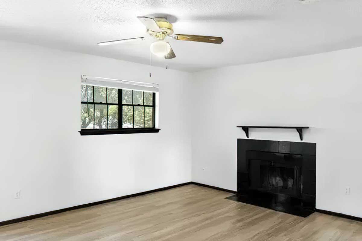 A bright, empty room featuring a ceiling fan, a large window with black trim, and a sleek black fireplace on one wall. The floor is hardwood, and the walls are painted white, creating a clean and spacious atmosphere.