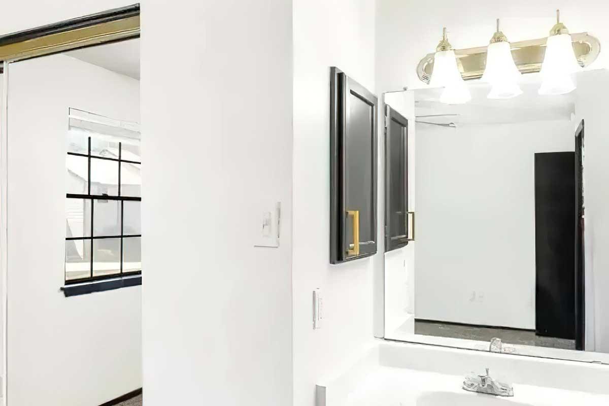 A bright, modern bathroom featuring a white sink and a large mirror with three light fixtures above. On the left, a partially open door leads to another room, and a window is visible, providing natural light. The walls are predominantly white, with a touch of black in the cabinetry.
