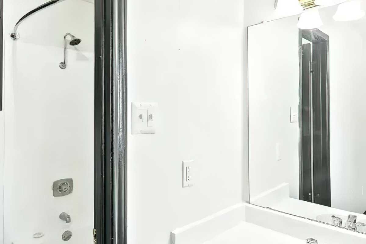 A bright bathroom featuring a walk-in shower with a glass door on the left side. Opposite the shower is a white sink with a modern faucet, beneath a large mirror. The walls are painted white, and the overall design is clean and minimalist.