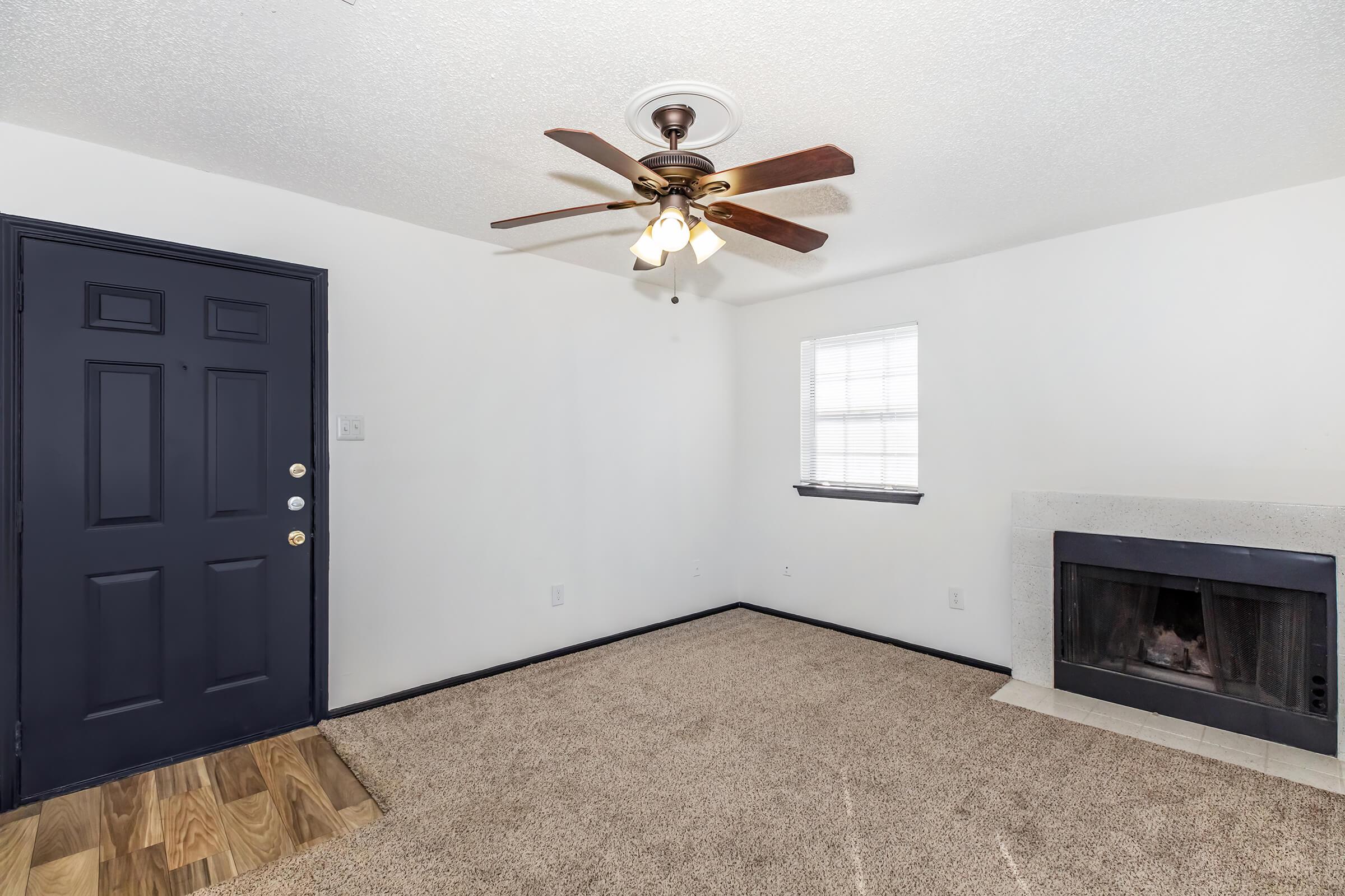 A small living room featuring a dark blue front door, a ceiling fan with lights, a window with blinds, and a fireplace with a stone surround. The floor is carpeted in a light beige color, and the walls are painted white.