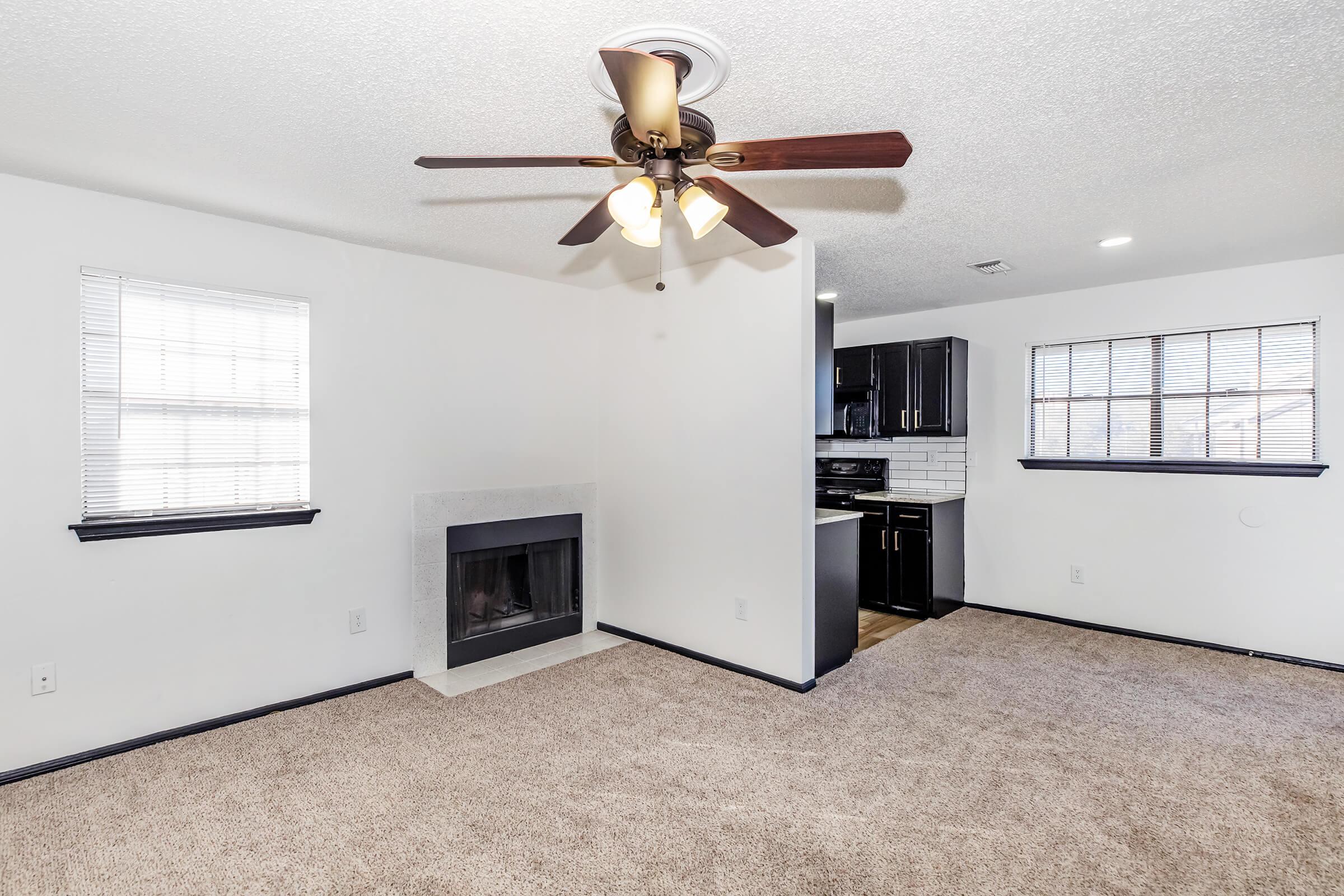 A cozy living area featuring beige carpet, a ceiling fan with lights, and a decorative fireplace. The space includes large windows with white blinds, providing natural light. A modern kitchen with dark cabinetry is visible in the background, creating an inviting and functional atmosphere.