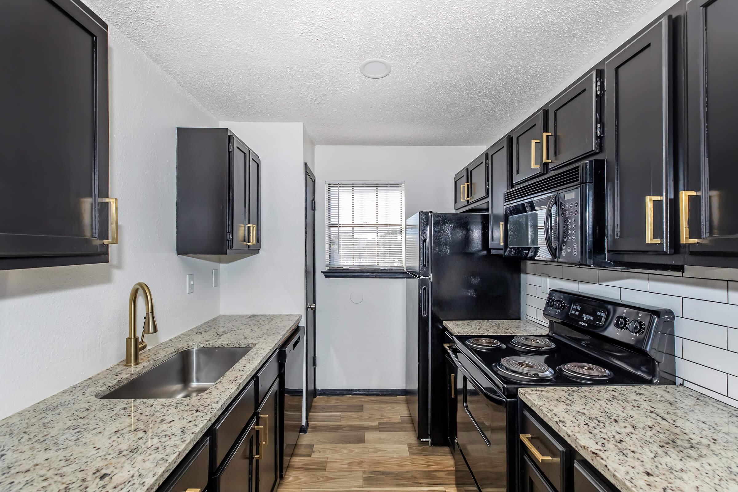 A modern kitchen featuring black cabinets with gold hardware, granite countertops, and stainless steel appliances. The layout includes a sink, stove, microwave, and refrigerator, with a window allowing natural light to brighten the space. The flooring is wood-like, adding warmth to the sleek design.