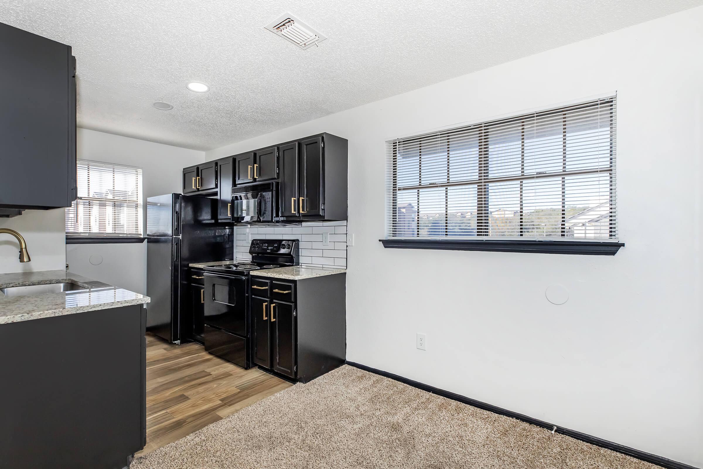 Modern kitchen featuring black cabinetry, a stainless steel stove and microwave, and white tile backsplash. The space includes a granite countertop and a window with blinds, allowing natural light to fill the area. The flooring is carpet, adding a cozy touch to the design.
