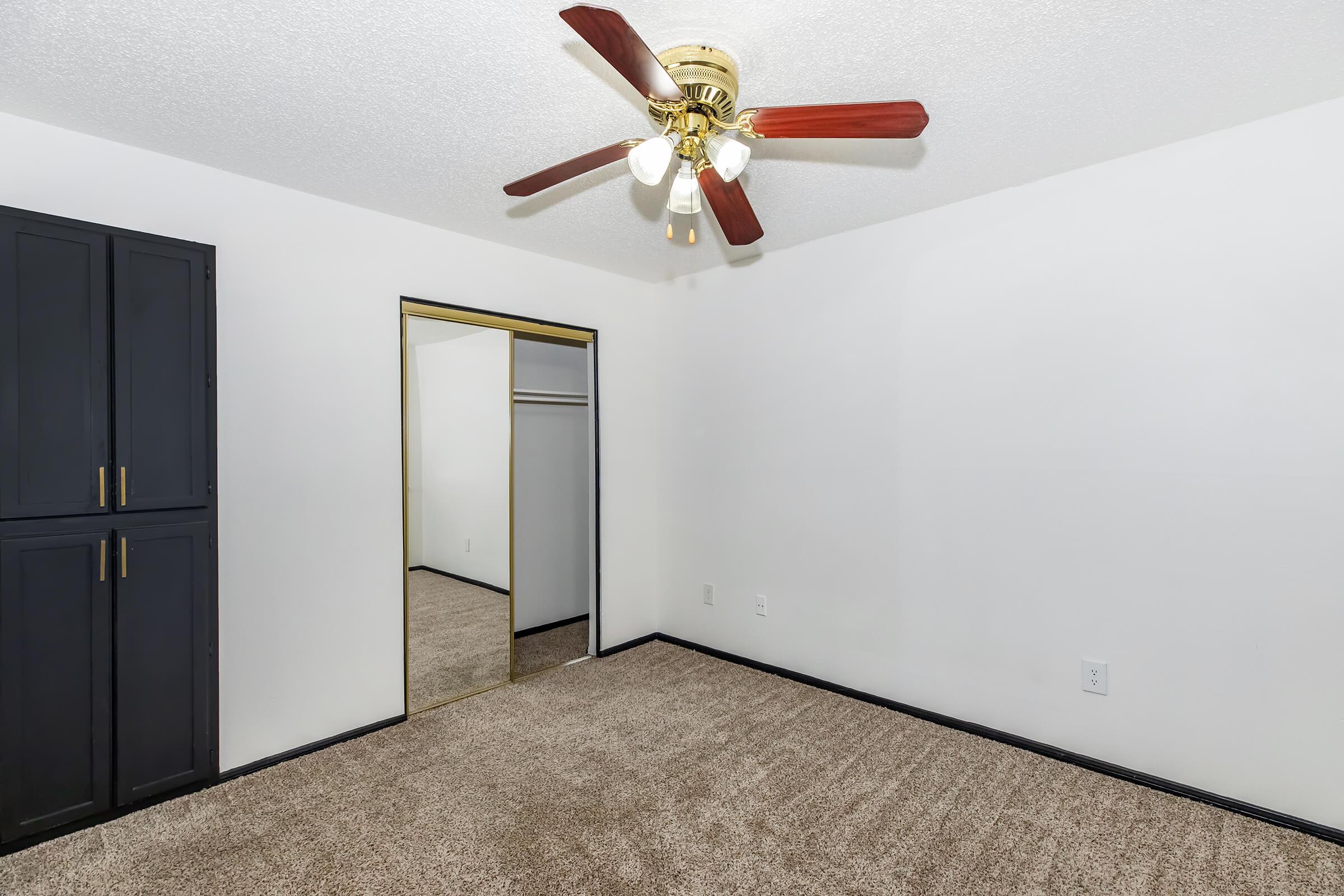 A clean, empty bedroom featuring beige carpet, a ceiling fan with five lights, and a dark wood closet with mirrored sliding doors. The walls are white, and there is a sense of spaciousness and simplicity in the design.