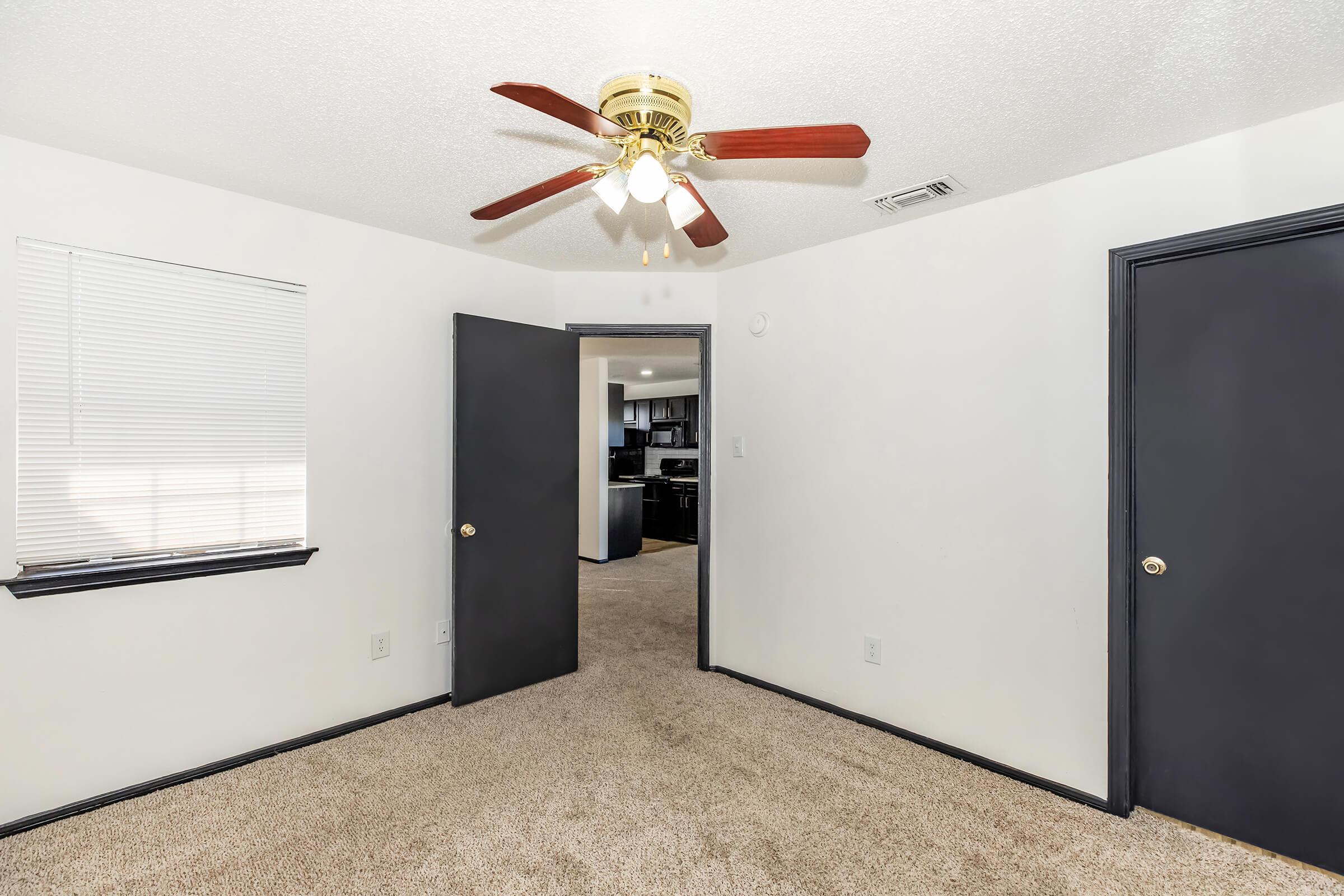 A well-lit room featuring beige carpet, a ceiling fan with wooden blades, and two doors leading to other areas. One door opens towards a kitchen or living area, while the other leads to the hallway. A window with blinds allows natural light to enter. The walls are painted white.