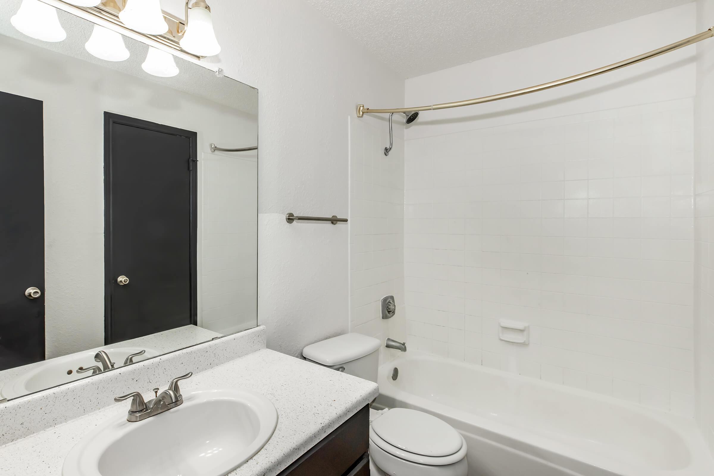 A clean and modern bathroom featuring a white bathtub and shower, a dark cabinet under a sink with a granite countertop, and a large mirror with three light fixtures above. The walls are painted white, and there's a dark door providing access to another room.