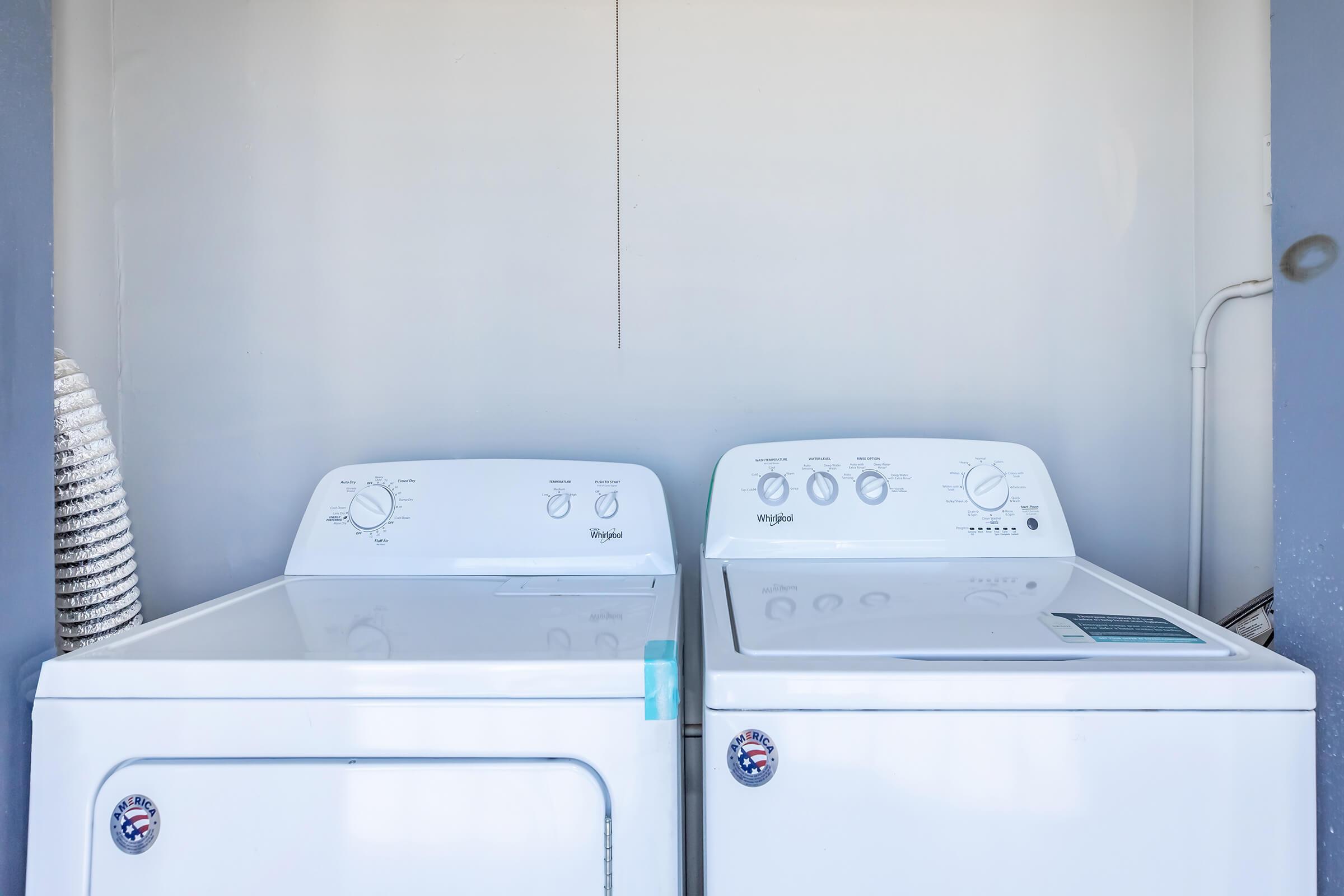 A side-by-side view of a white washing machine and dryer. Both appliances feature control knobs and dials on the front. The washing machine is on the right and the dryer on the left, with a vent hose visible on the left side. The appliances are set against a plain wall.