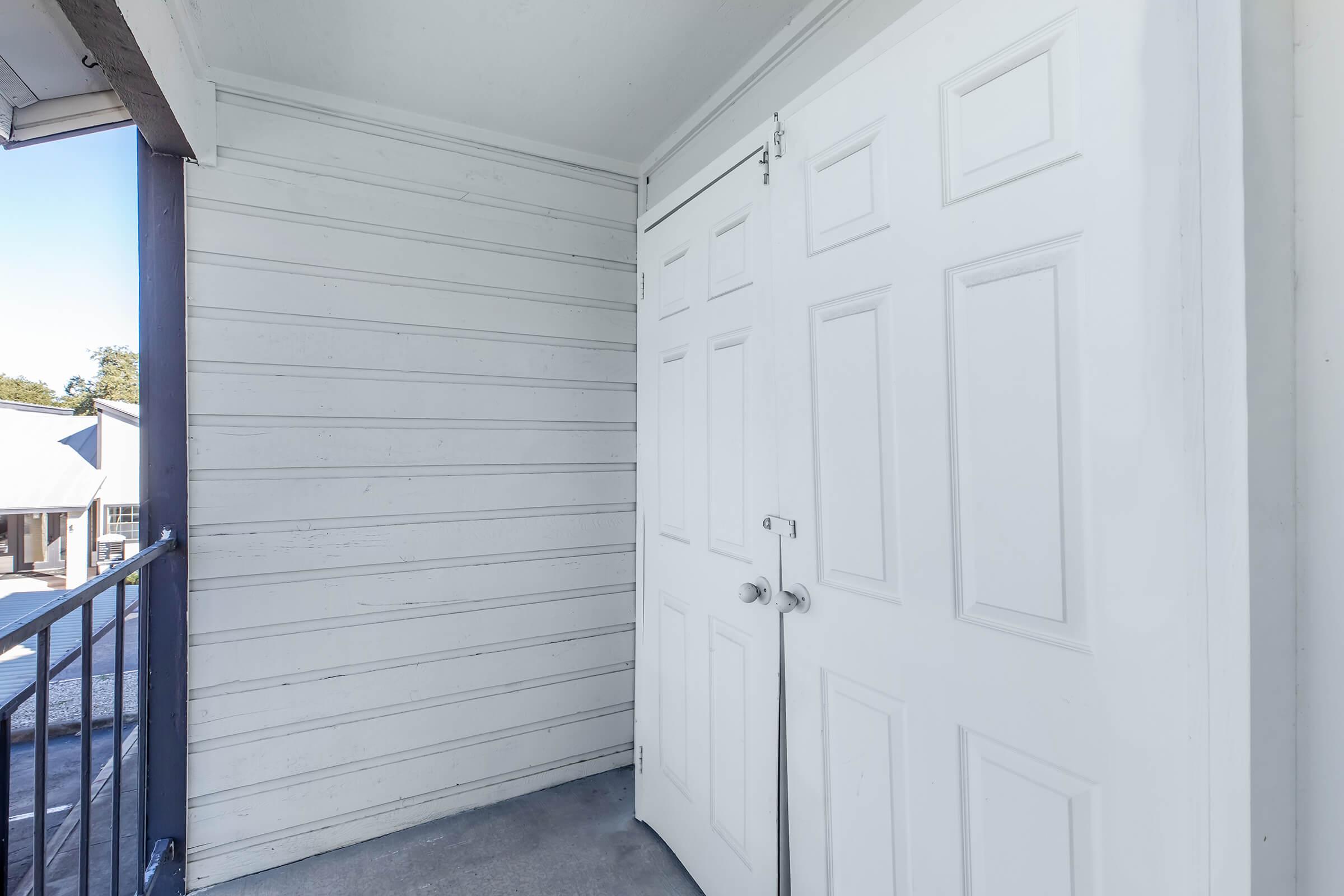 A white double door on a balcony with a railing. The walls are light-colored paneling, and the door is in an open position, revealing a small storage space. In the background, there's a glimpse of a building and a clear blue sky.