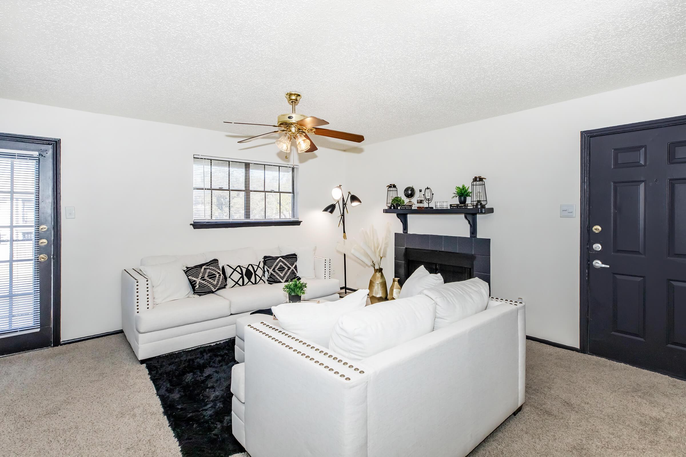 A cozy living room featuring two white sofas with decorative pillows, a black rug, a ceiling fan, and a small indoor plant. The room has two windows with black frames, a minimalist fireplace, and stylish decor on the mantle, creating a welcoming atmosphere.