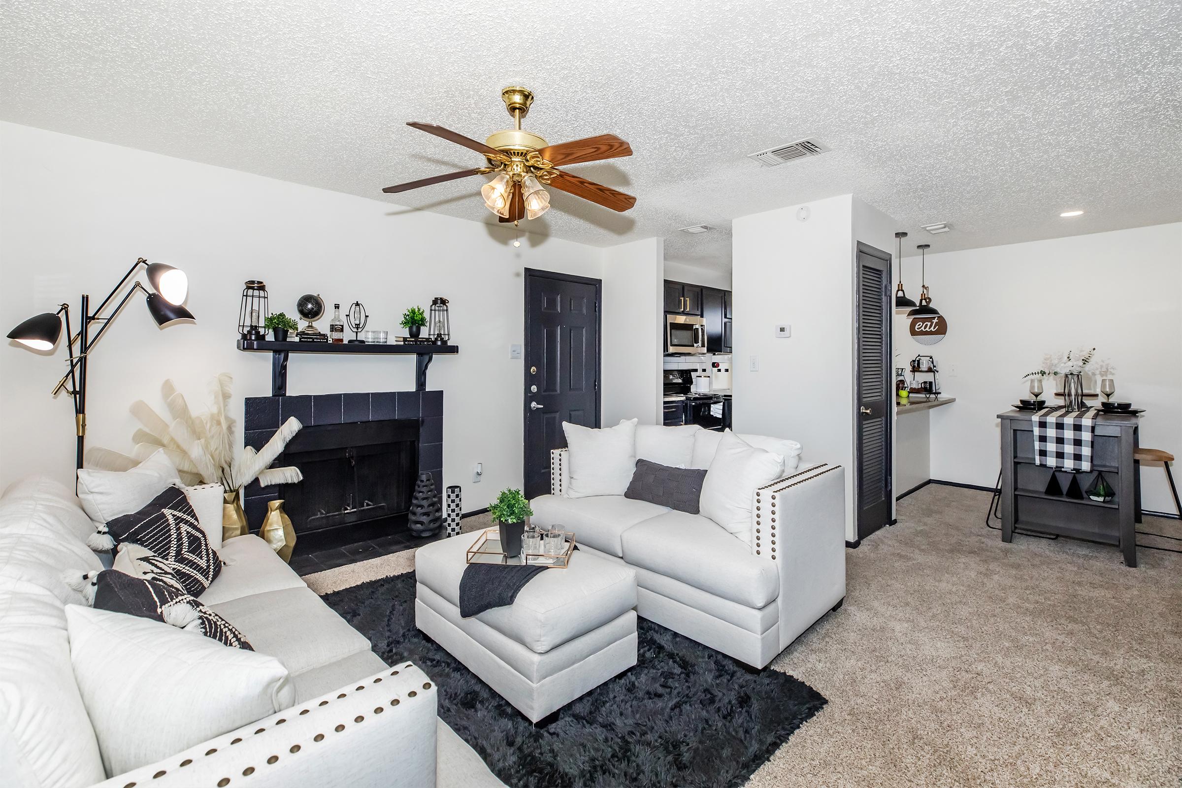 A cozy living room featuring a white sectional sofa and an armchair with decorative pillows, a black fireplace with shelves, a ceiling fan, and a small dining area with a bar cart. The space is adorned with plants and stylish decor against a neutral-colored wall and plush carpet.