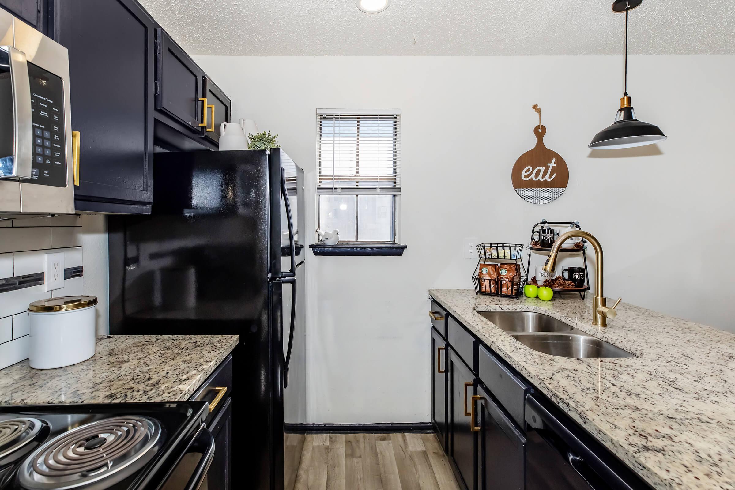 A modern kitchen featuring dark cabinetry, a black refrigerator, and a double sink with a gold faucet. The counter is topped with speckled granite, and a decorative "eat" sign hangs on the wall. A window allows natural light, and a bowl of green apples is visible on the counter.