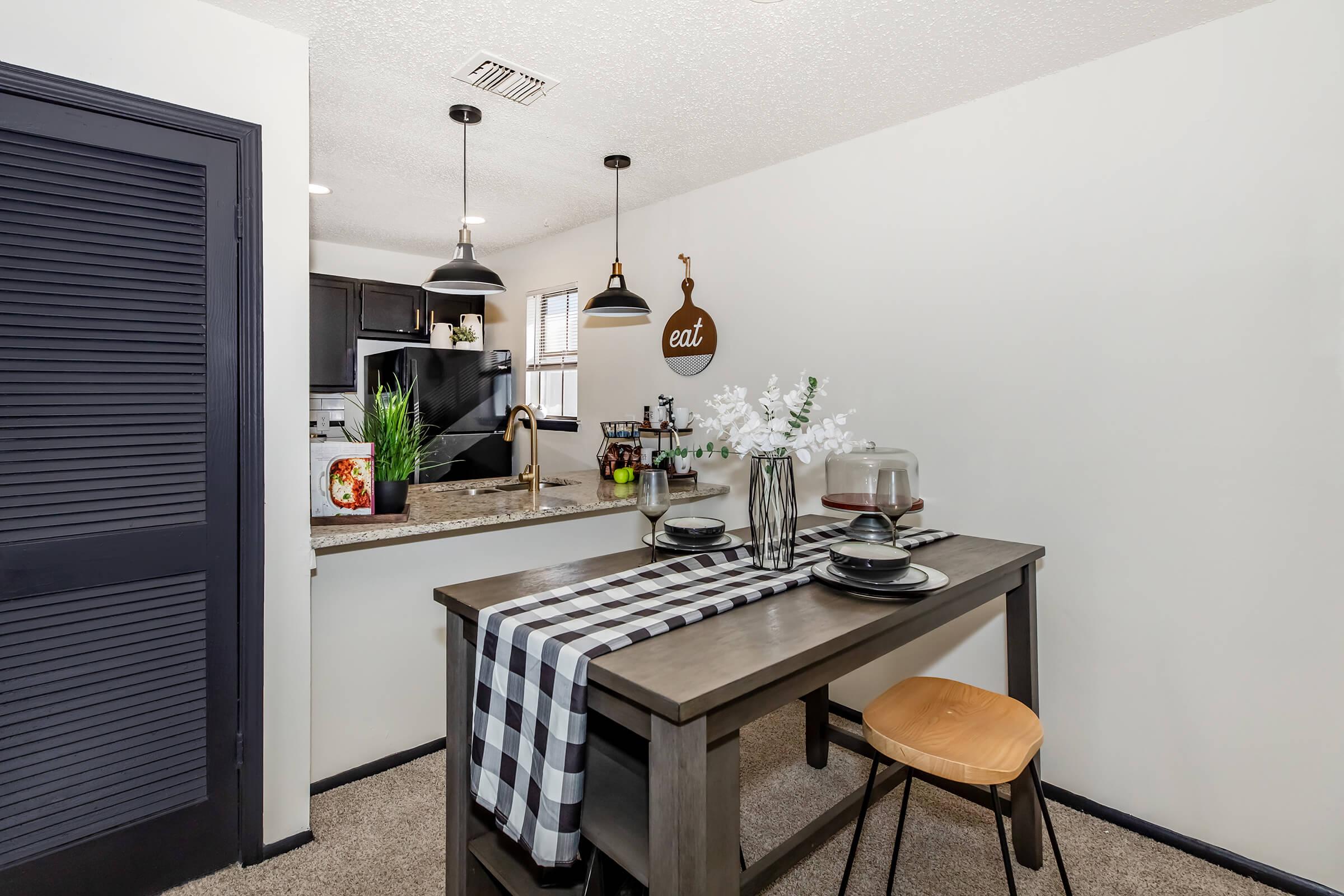 A modern dining area featuring a wooden table set for two with black and white tableware, a checkered table runner, and fresh flowers in a vase. In the background, a sleek kitchen is visible with dark cabinetry. Pendant lights hang overhead, creating a cozy atmosphere.