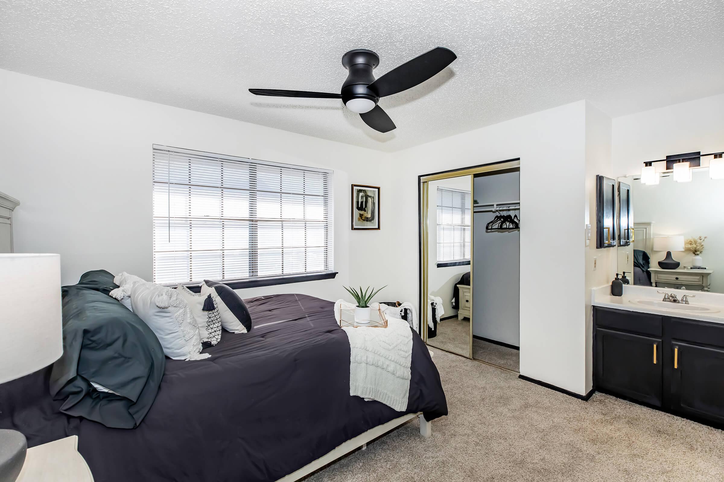 A well-decorated bedroom featuring a bed with dark bedding, a ceiling fan, and natural light streaming through windows. There’s a closet with hanging clothes and a bathroom area visible. The decor includes a lamp and plants, creating a cozy and inviting atmosphere.