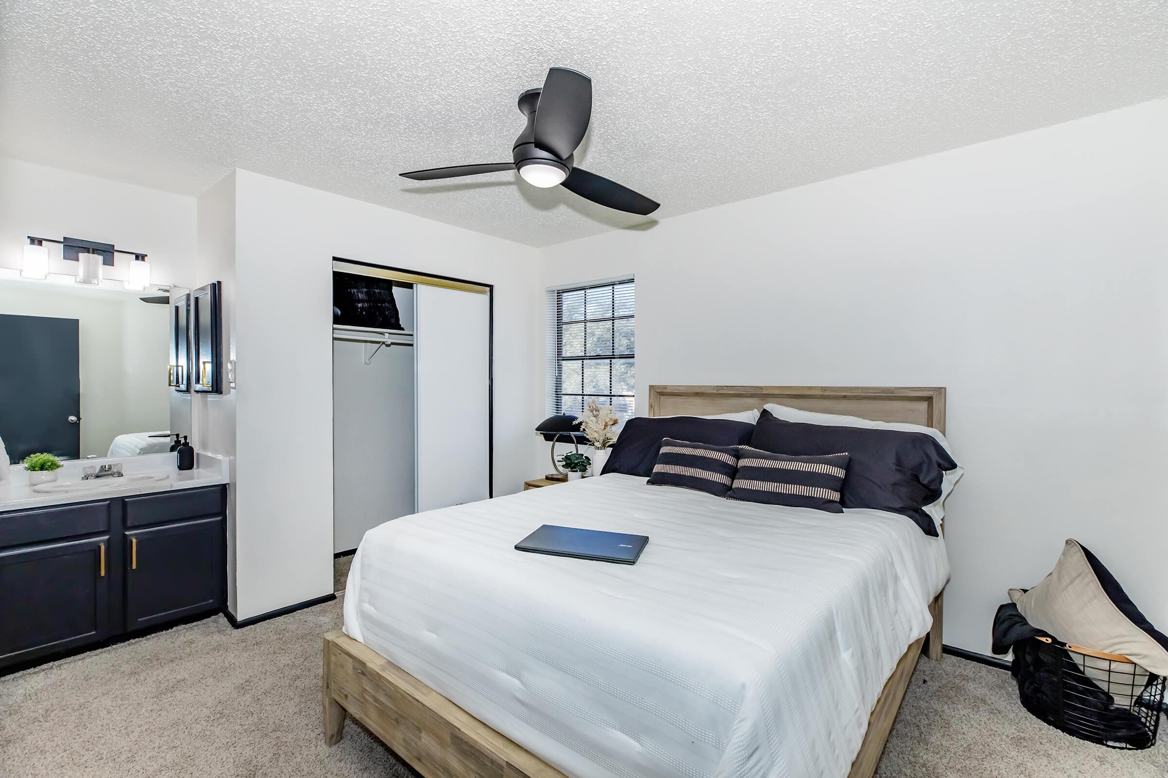 A cozy bedroom featuring a bed with a light-colored duvet, two decorative pillows, and a laptop resting on it. The room has a modern ceiling fan, a nightstand with a lamp, and a closet with sliding doors. Natural light streams through a window, illuminating the neutral-colored walls and carpet.