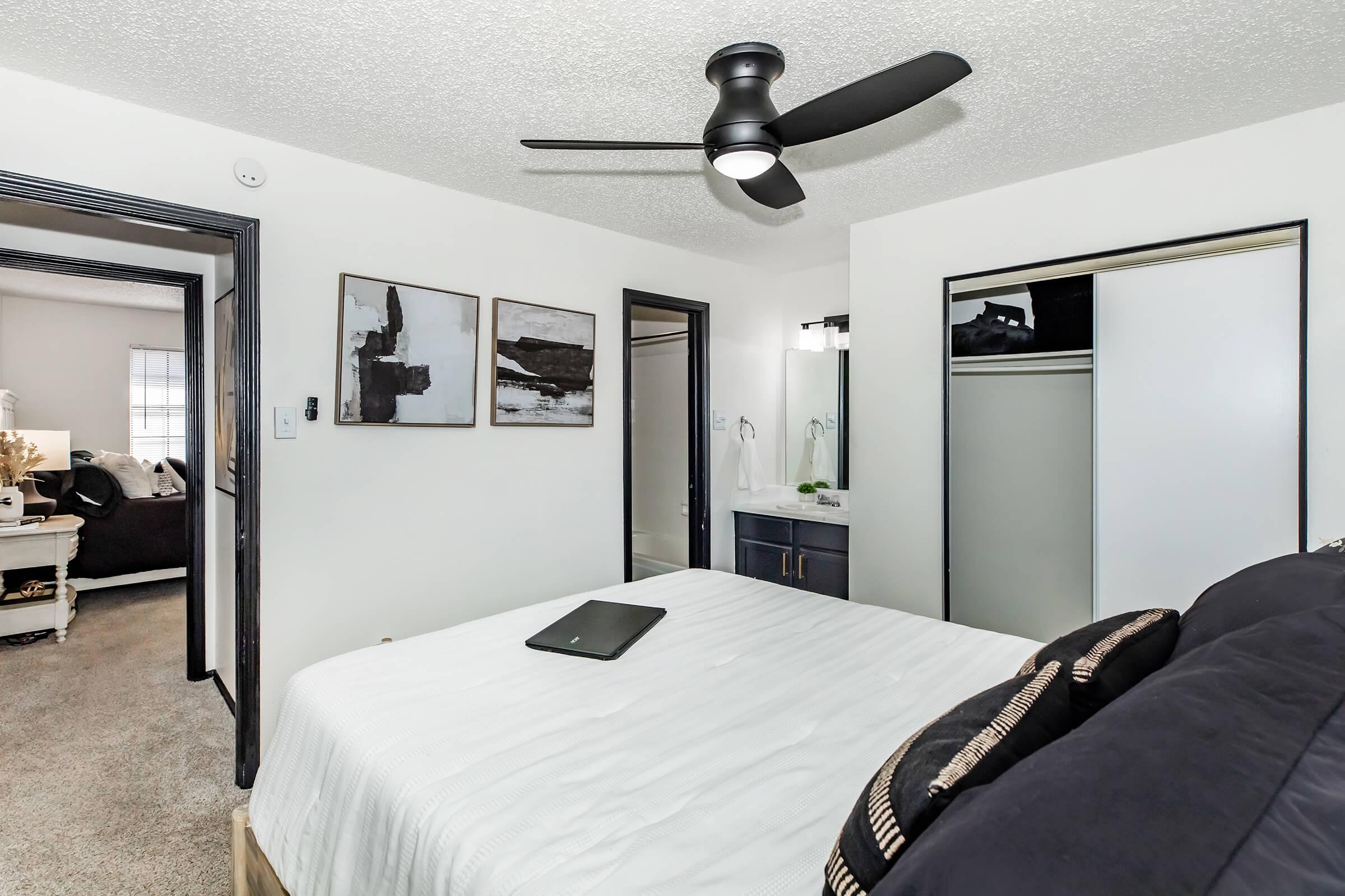 A cozy bedroom featuring a bed with a black and white bedding set, a ceiling fan, and art pieces on the wall. Two doorways lead to other rooms, and a bath area is partially visible. The decor is modern and minimalist, with a neutral color palette and soft lighting.